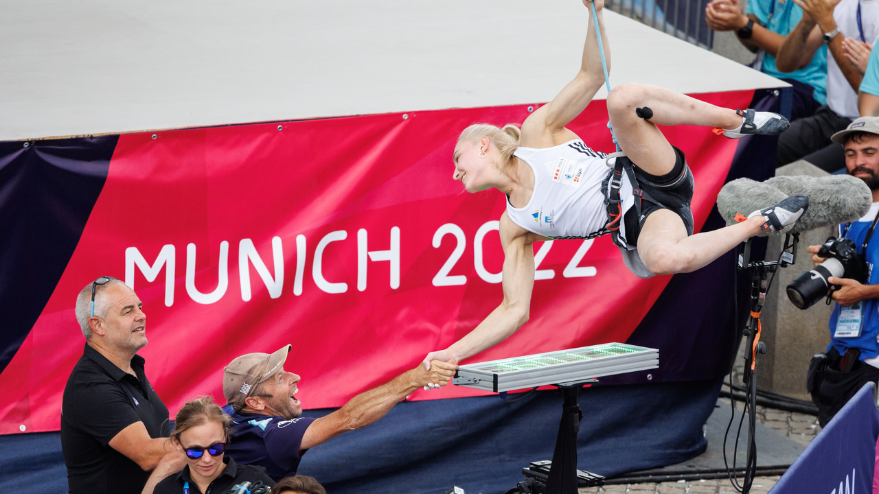 Eine Kletterin feiert ihren Erfolg bei den European Championships 2022 in München, während sie am Zielgriff hängt und einer Person aus dem Team die Hand gibt.