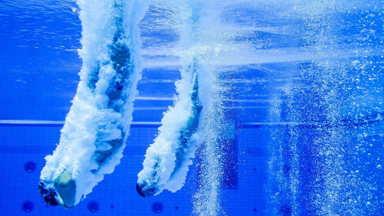 Zwei Wasserspringer tauchen gleichzeitig ins Becken ein, Blasen steigen auf.