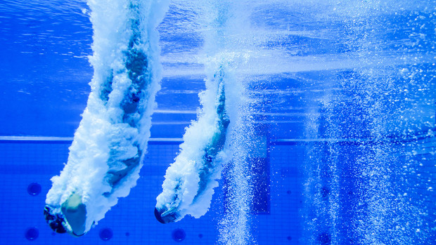 Zwei Wasserspringer tauchen gleichzeitig ins Becken ein, Blasen steigen auf.