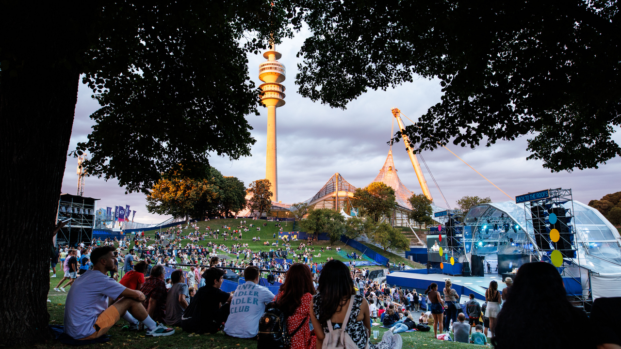 Festivalbesucher sitzen auf einer Wiese mit Blick auf Bühne und Fernsehturm.