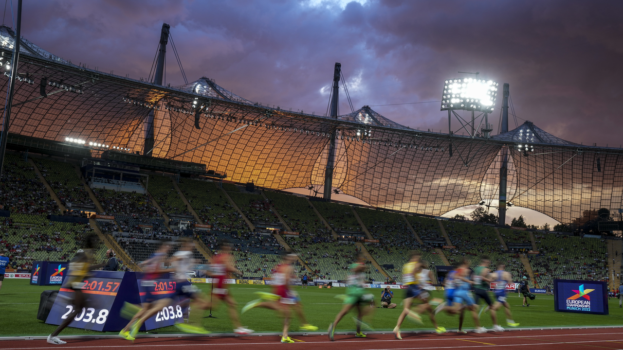 Läufer rennen im Stadion bei Sonnenuntergang unter beleuchtetem Zeltdach vorbei.