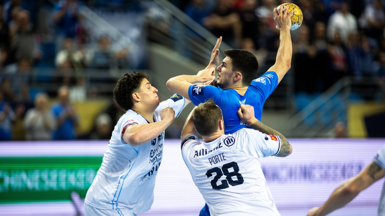 Handballspieler im blauen Trikot springt zum Wurf, zwei Gegenspieler in Weiß blocken ihn.