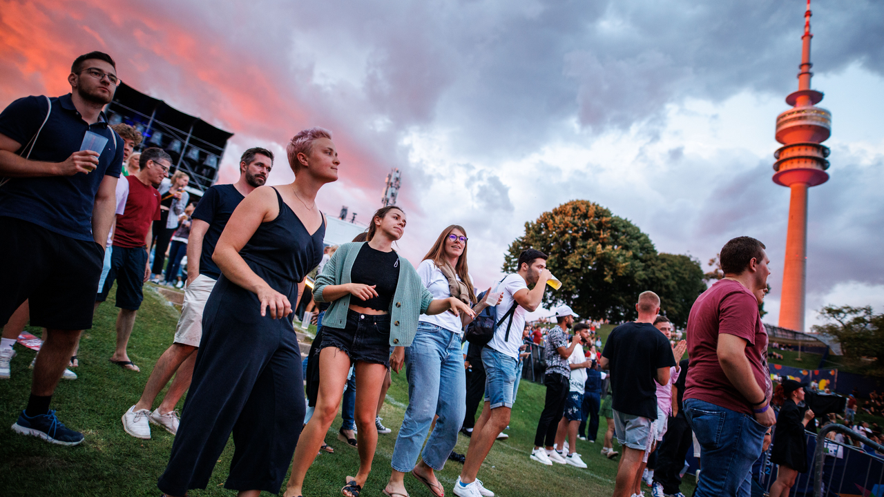 Festivalbesucherinnen und -besucher tanzen und genießen den Abend im Olympiapark München während der European Championships 2022. Im Hintergrund ist der Olympiaturm vor einem farbenreichen Sonnenuntergang zu sehen.