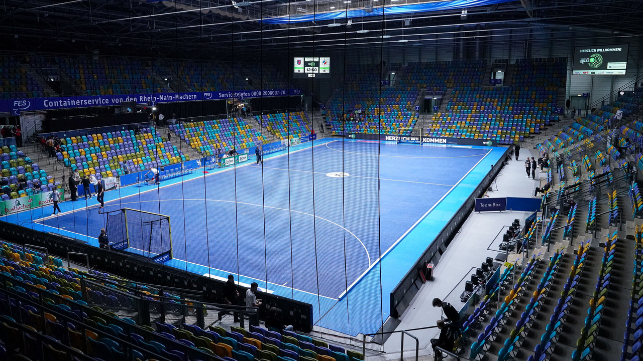 Leere bunte Tribünen in einer großen Halle mit blauem Hallenhockeyfeld.