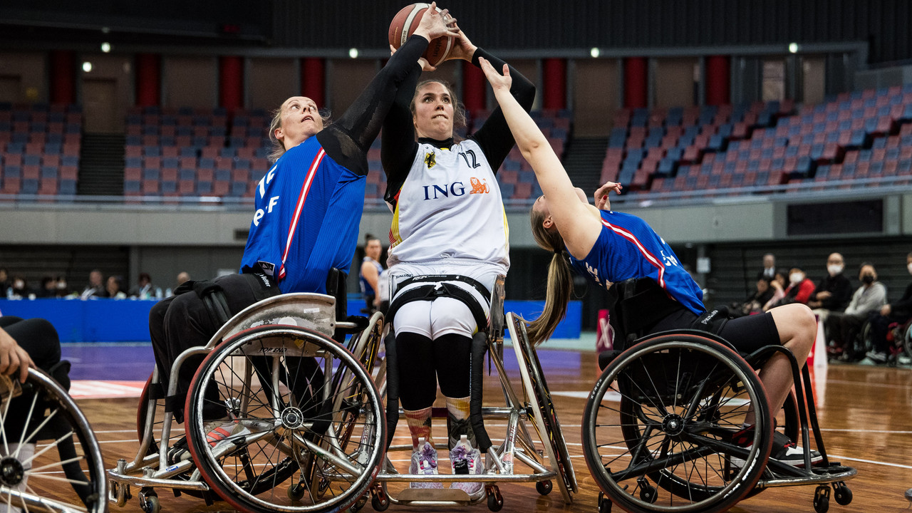 Rollstuhlbasketballspielerin im weißen Trikot hält den Ball hoch, zwei Gegnerinnen versuchen zu blocken.