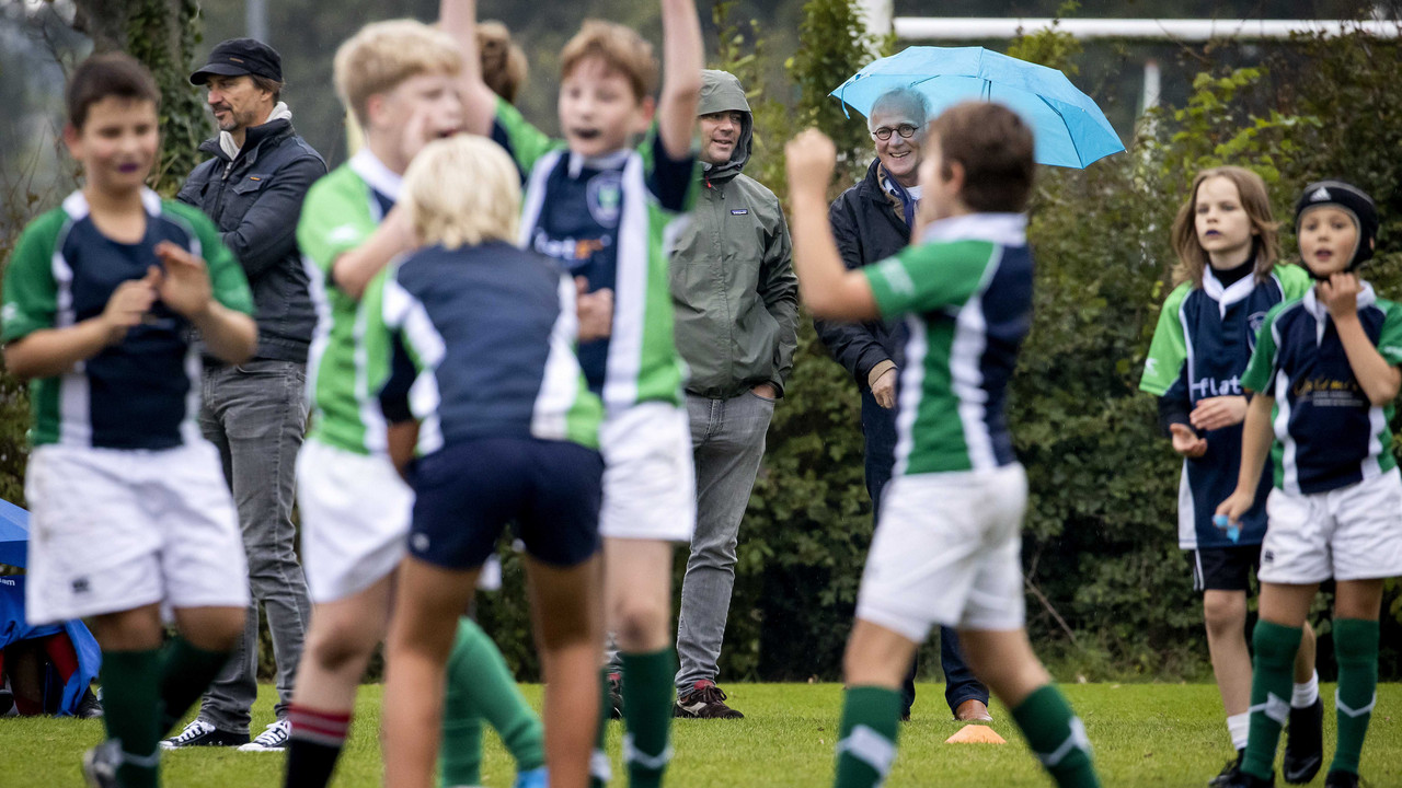 Kinder spielen Rugby auf einem Rasenplatz, Erwachsene schauen im Hintergrund zu.