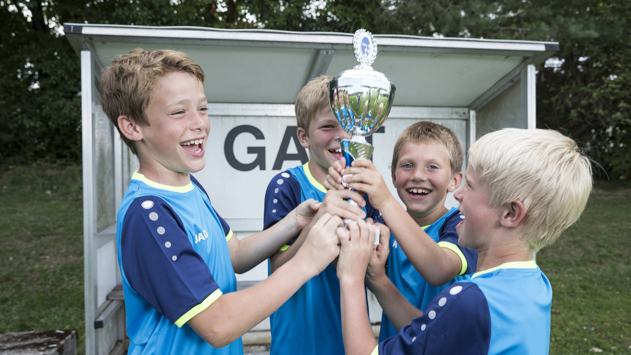 Vier Kinder im blauen Trikot jubeln und halten gemeinsam einen Pokal hoch.