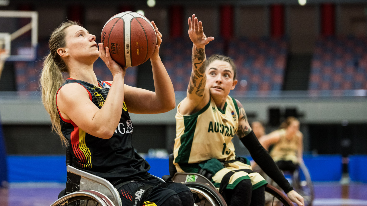 Rollstuhlbasketballspielerin im schwarzen Trikot wirft den Ball, Gegenspielerin in Gelb verteidigt.