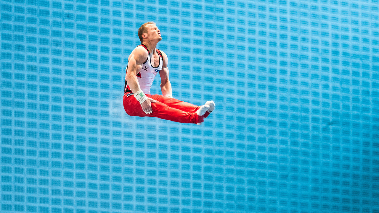 Turner in roter Hose springt hoch über dem Reck und zieht die Beine an.