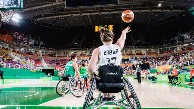 Rollstuhlbasketballspielerin wirft den Ball in einer großen Halle, Gegnerinnen schauen zu.