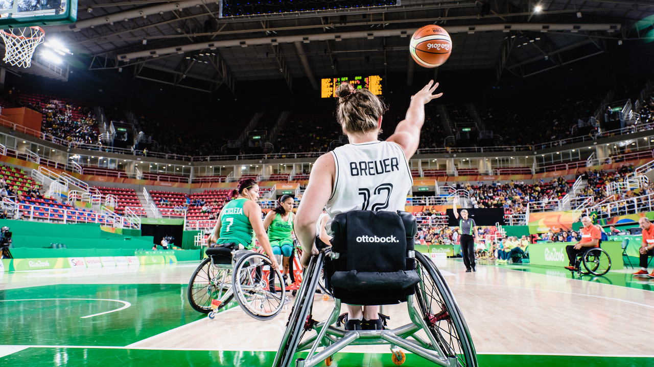 Rollstuhlbasketballspielerin wirft den Ball in einer großen Halle, Gegnerinnen schauen zu.