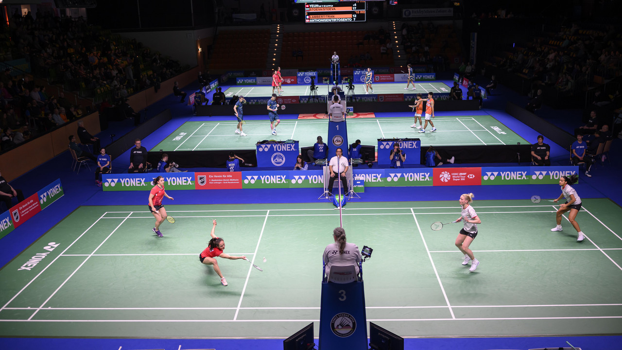 Badminton-Doppel spielen auf mehreren Feldern in einer Halle vor Publikum.